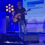 Na scenie stoi Wojciech Cugowski, który śpiewa i gra na gitarze. W tle ekran, na którym wyświetla się napis 